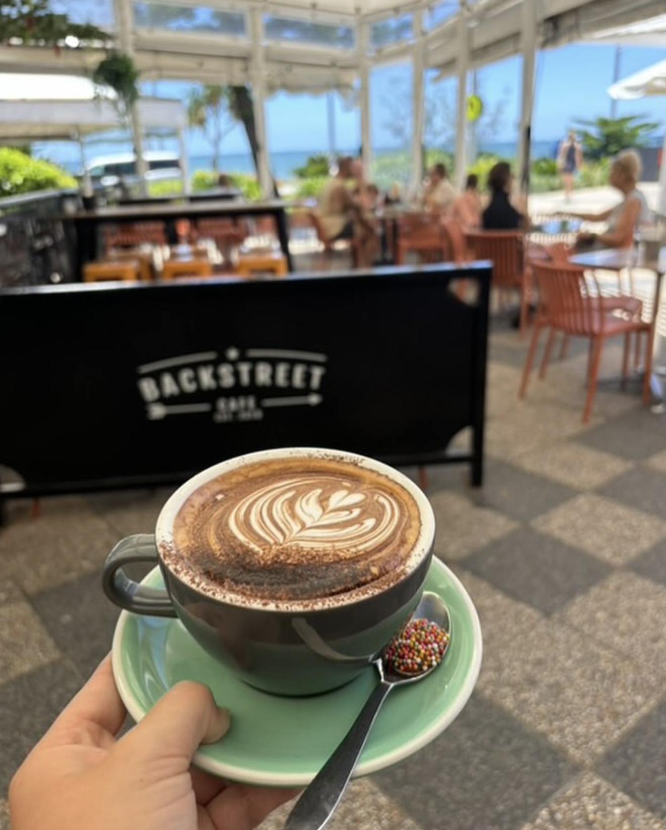 A mocha with latte art held up on the Backstreet Cafe terrace, with beachfront tables and the ocean behind