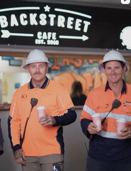 Two tradesmen in hi-vis leaving Backstreet Cafe with takeaway coffees, under the lit Backstreet Cafe sign